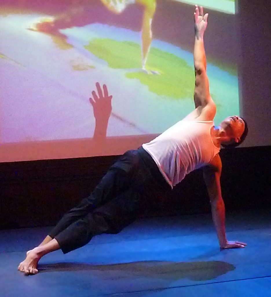Photo of Learie Mc Nicolls dancing, supporting himself with one arm, in front of projected image. 